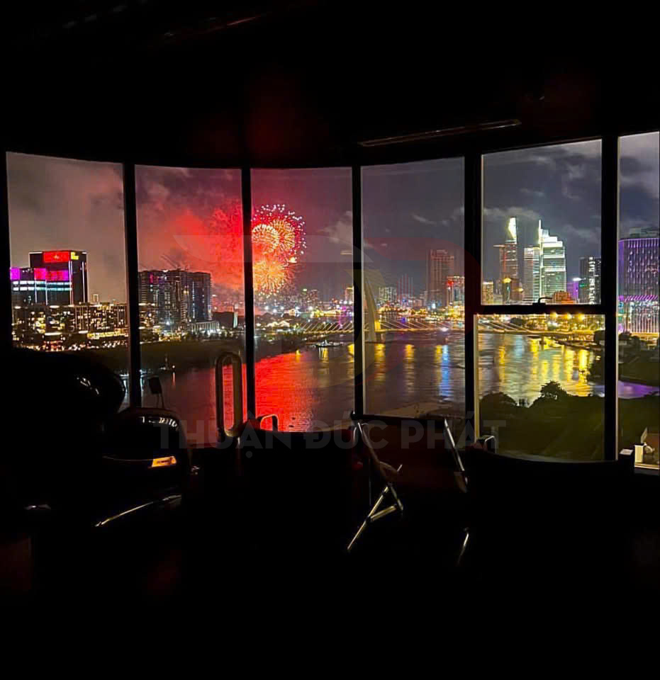 Phòng làm việc với cửa kính lớn nhìn ra sông và pháo hoa rực rỡ trên nền skyline thành phố ban đêm